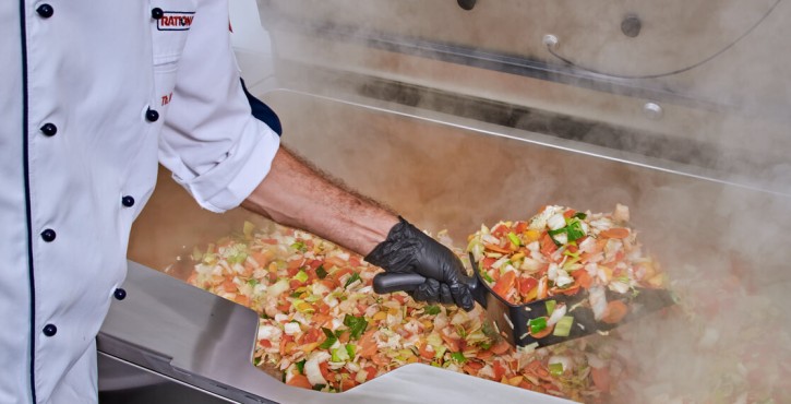 A RATIONAL chef can be seen with a black glove on using a scoop to lift out vegetable stir fry from an iVario Pro L which is steaming with freshly cooked vegetables.