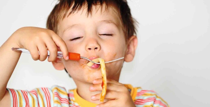 A young child in a multicoloured stripey top eats a stand of tomato pasta off a fork, enjoying it with his eyes closed and sauce all round his mouth.