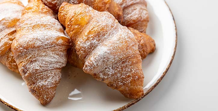 A pile of golden baked croissants sit on a white plate, dusted with icing sugar.