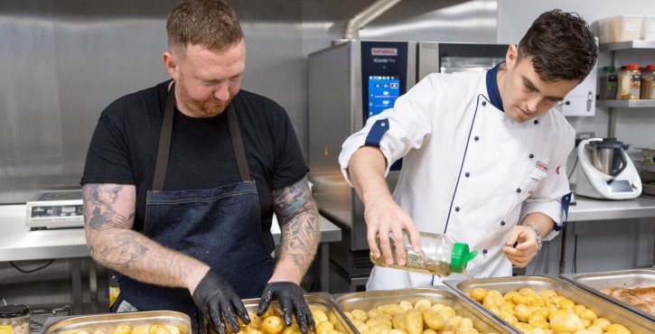 A chef in a white jacket sprinkles herbs onto oven trays of potatoes whilst another chef in a black t shirt and blue denim apron rubs oil over the potatoes. The both stand in front the RATIONAL iCombi Pro in the kitchen.