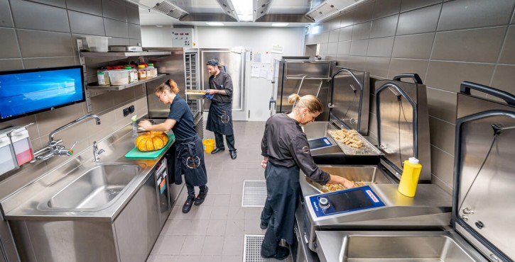 A zoomed out shot of a small working kitchen with 2 female chefs and 1 male chef, all in dark uniforms all preparing different food at different stations. 