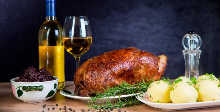 A roast duck sits on the table surrounded by delicious potato dumplings, braised red cabbage and white wine.