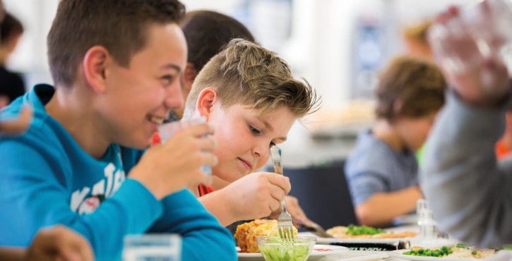 Children eating school lunches, one boy is eating cucumber from his lunch tray with a fork