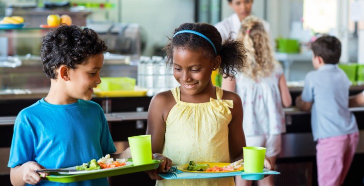 A young black school girl and young Hispanic school boy look at each other’s lunch trays as they carry them across the school cafeteria