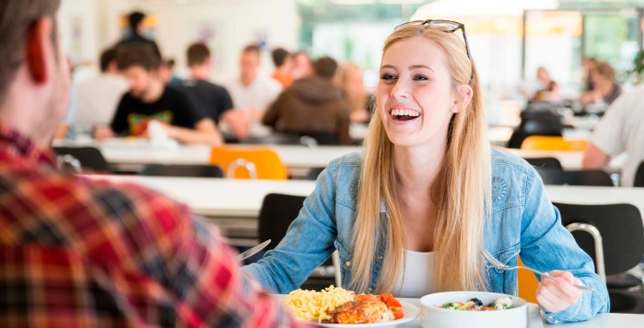 A blonde female in a denim blue jacket laughs at a fellow student across the cafeteria table. A tray of chicken, potatoes and salad sits on the table in front of her.