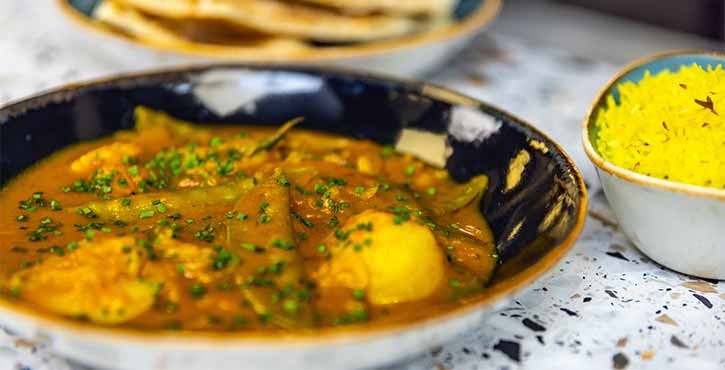 A bowl of creamy vegan curry sits on the countertop, a rich orange sauce with potatoes and mangetout. Some golden pilau rice and freshly baked naan sit in the background.