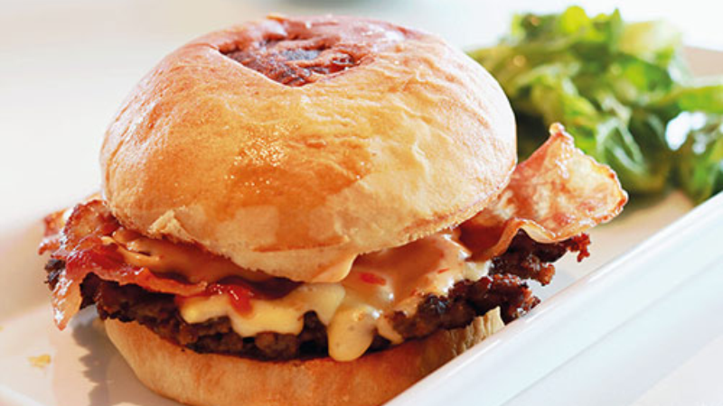 A burger with crispy bacon, melted cheese, and sauces on a bun, served with a side salad on a white plate.