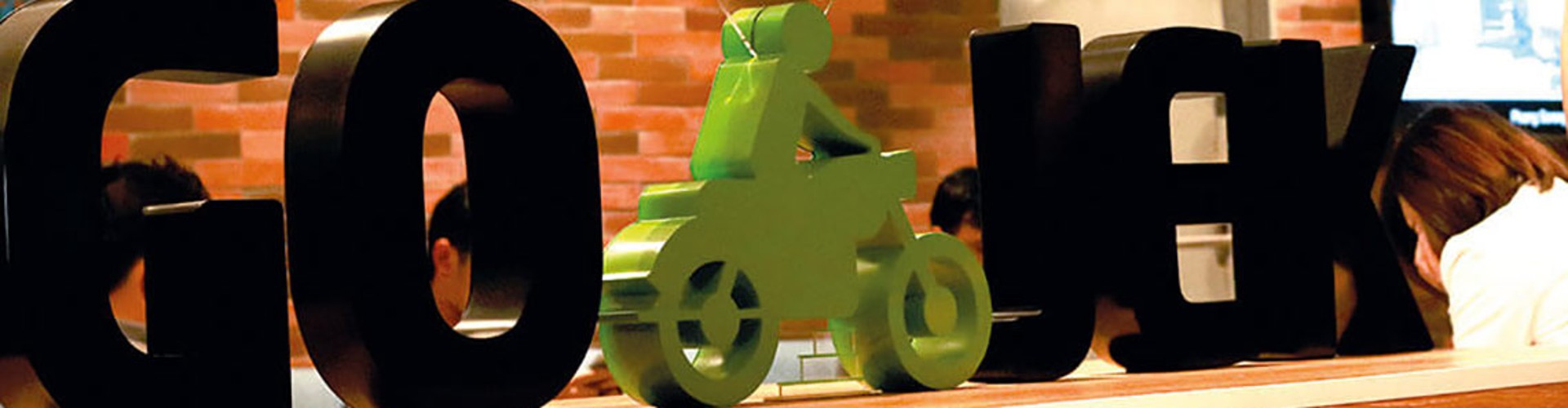 Logo of Go-Cafe with a toy motorcycle placed on it, seen on a blurry background of people sitting at a table.