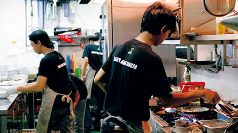 Two chefs working in a busy kitchen at Go-Cafe, one prepping ingredients and the other cooking on a stove.
