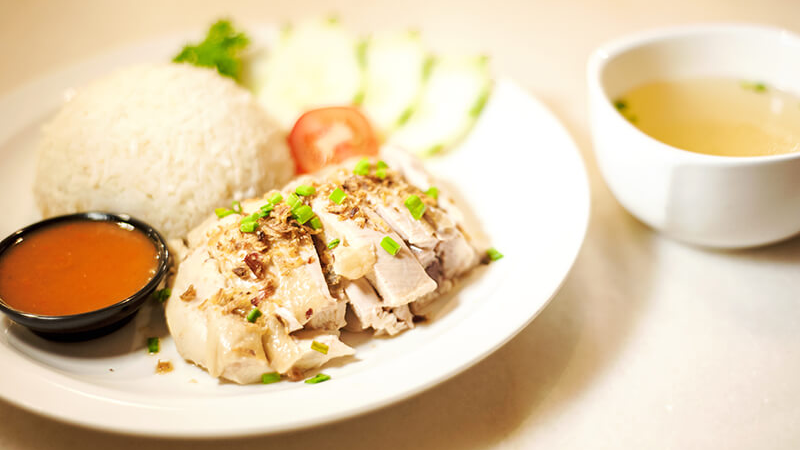 A plate of Hainanese chicken rice with slices of steamed chicken, white rice, and garnishes of cucumber and tomato, accompanied by a bowl of soup and a side of chili sauce.