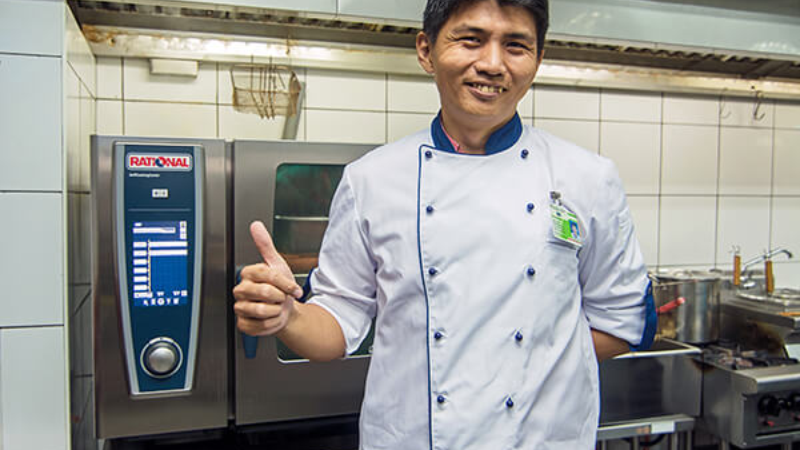 A chef in a white uniform smiling and giving a thumbs up in a commercial kitchen next to a Rational oven.