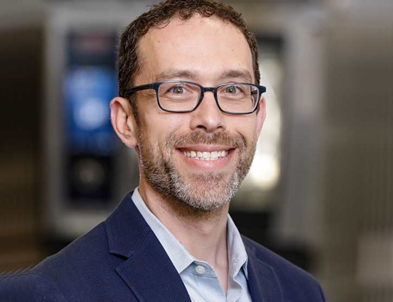 Portrait of a smiling individual wearing glasses, a blue blazer, and a light blue collared shirt, set against a softly blurred office background.