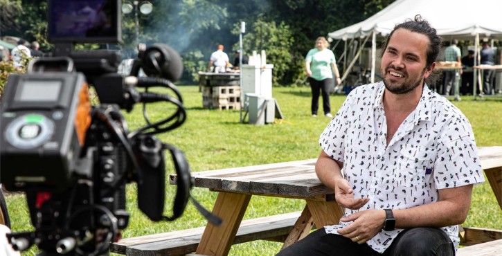 Smiling male chef in a patterned short-sleeve shirt sits at a picnic table outdoors during a filmed culinary event interview
