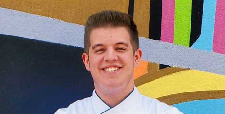 Young smiling chef in white chef‘s coat with arms crossed standing in front of a colorful abstract mural