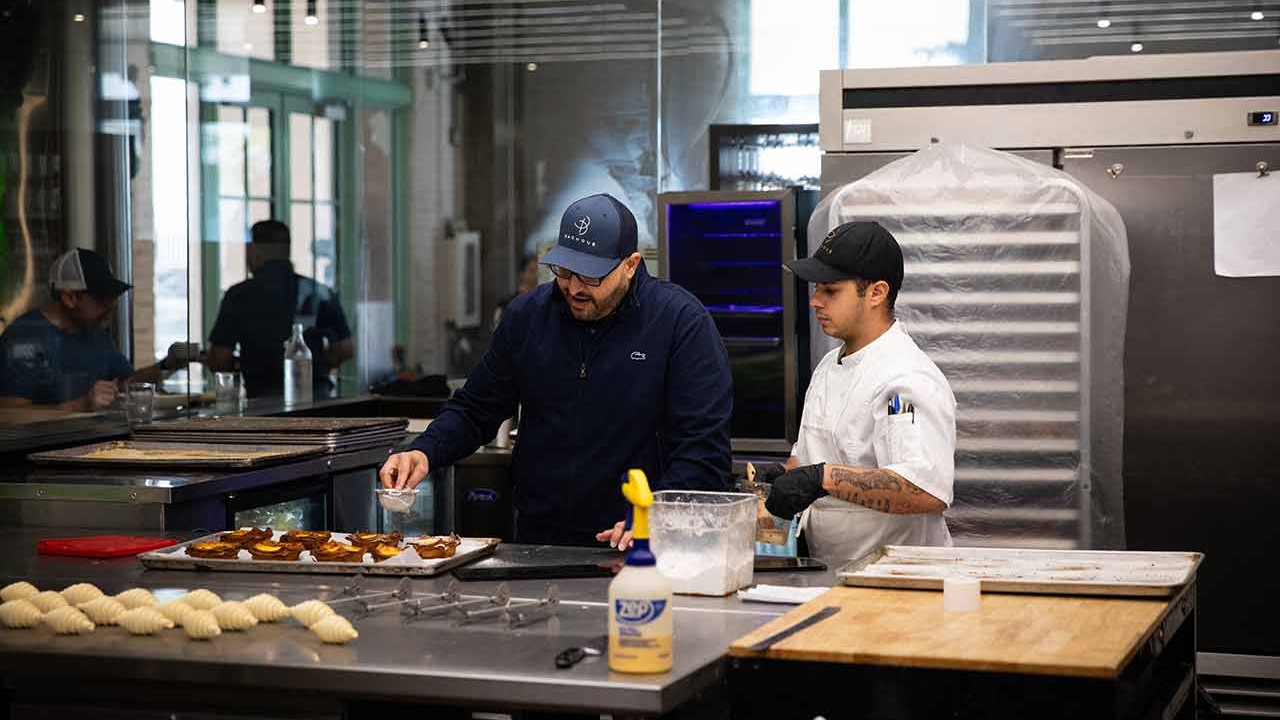 Two chefs collaborating in a modern kitchen, one sprinkling seasoning on a tray of food while the other assists.