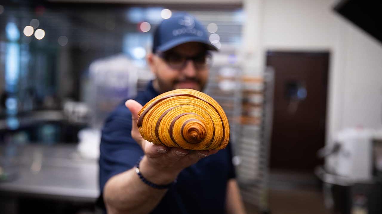 Chef holding a beautifully crafted croissant in a kitchen setting, with baking equipment in the background.
