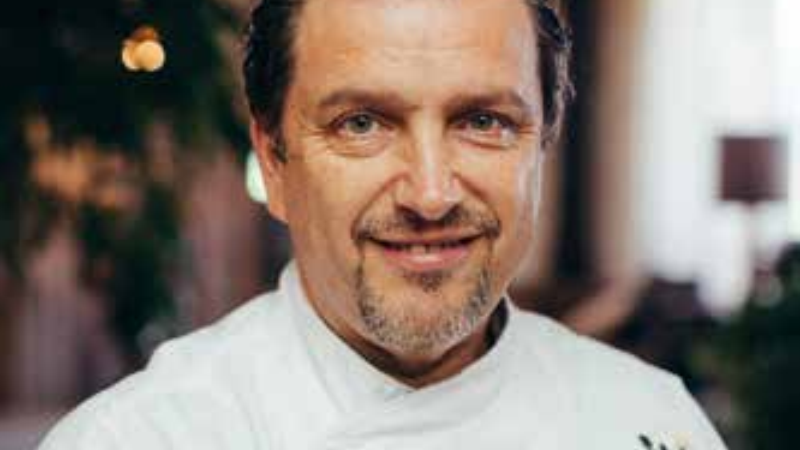 Chef smiling in a kitchen setting, wearing a white chef‘s coat.