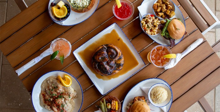 Overhead view of seafood dishes, burgers, cocktails and sides served on a wooden table at Coastal Orange Beach.