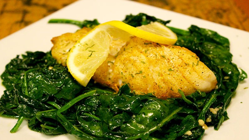 A plate of grilled fish fillet served on a bed of sautéed spinach, garnished with lemon slices and herbs.