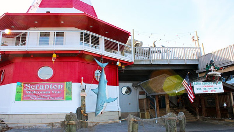The exterior view of Cooper‘s Seafood House, featuring a lighthouse-style building with a large fish sculpture, welcoming banners, and a sign for seafood market hours.