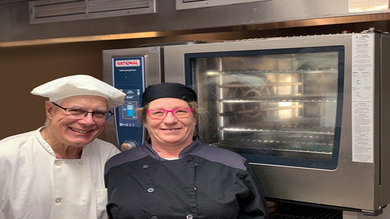 Two chefs, one in a white chef‘s hat and the other in a black chef‘s uniform, pose together in front of a Rational oven, smiling at the camera.