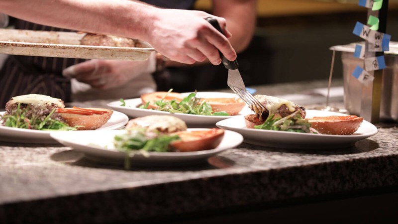 A chef plating burgers topped with melted cheese, served on plates with fresh salad greens on the side.