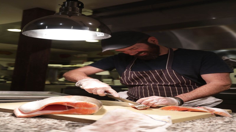 A chef skillfully filleting a large salmon on a cutting board in a busy kitchen, smiling as he works.