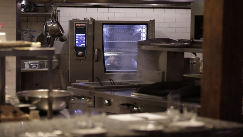 A professional kitchen featuring a Rational combi-steamer, surrounded by stainless steel equipment and cooking utensils, with steam rising from the oven.