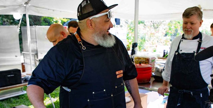 A chef with a beard and sunglasses cooks outdoors, surrounded by colleagues and kitchen equipment.