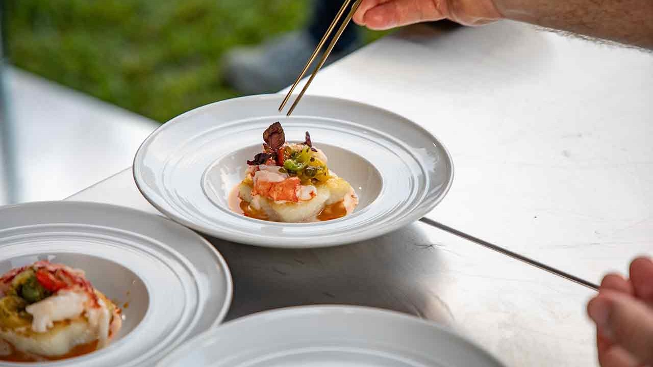 A close-up of a dish being garnished with chopsticks, featuring seafood and vegetables, with another plated dish visible in the foreground.