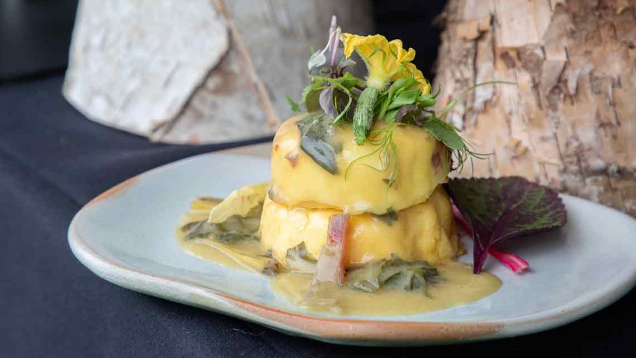 A plated dish featuring layers of food with a creamy sauce, garnished with fresh herbs and edible flowers, displayed on a rustic table.