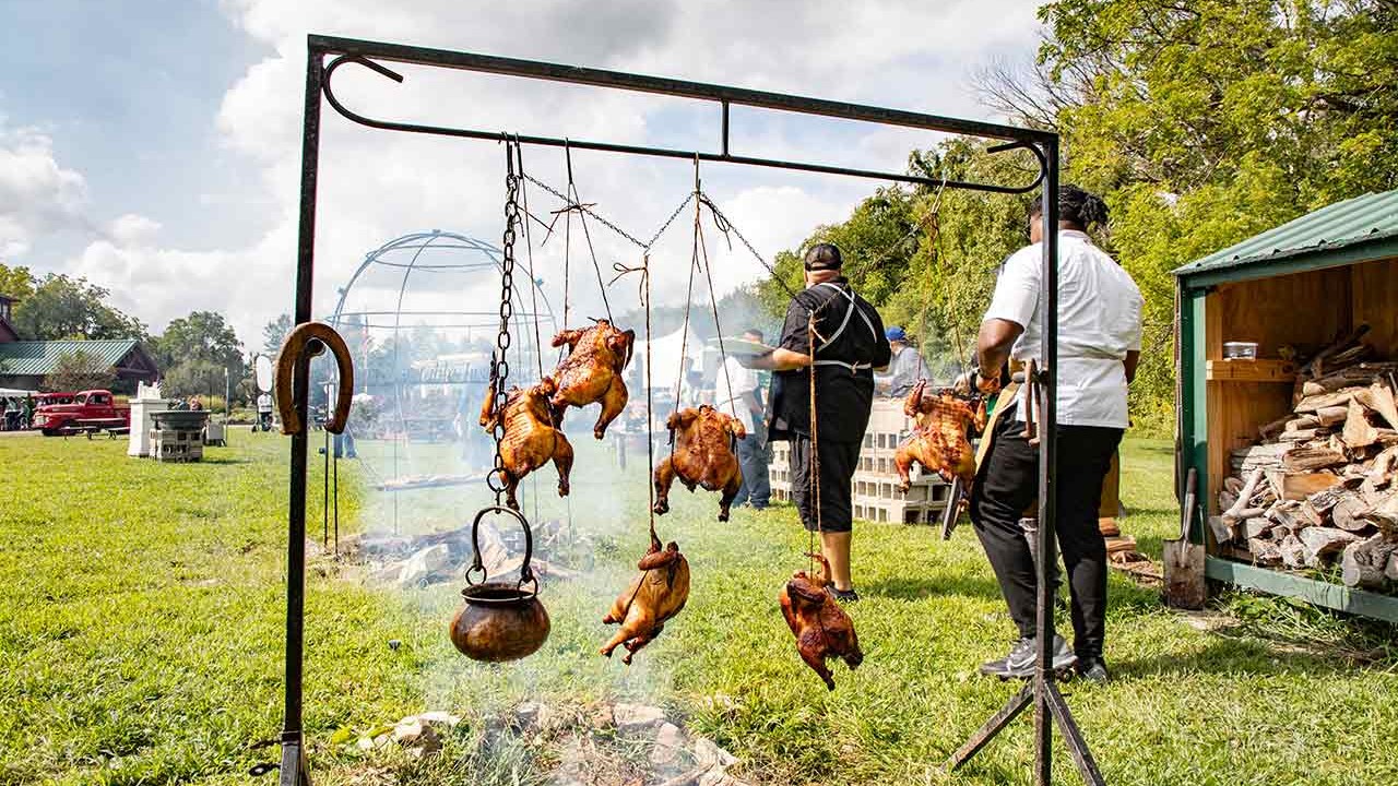 A scene of grilled chickens hanging over a fire pit, with chefs preparing food outdoors under a cloudy sky.