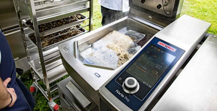 A close-up of a Rational iVario Pro cooking unit with steam rising, showing food being prepared inside, and a rack of prepared dishes in the background.