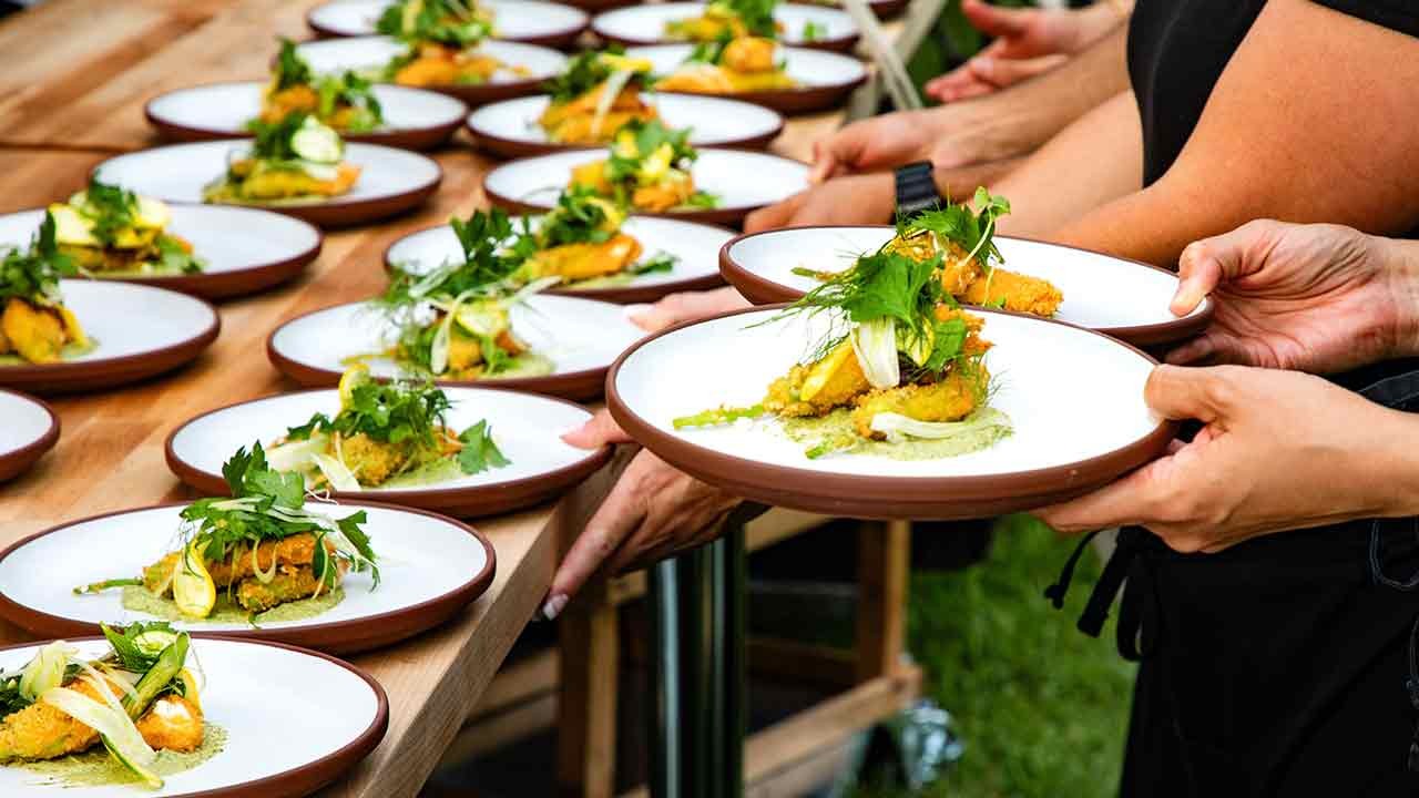 A vibrant outdoor scene showing several plates of food being served, with hands reaching out to take the dishes filled with garnished and colorful ingredients.