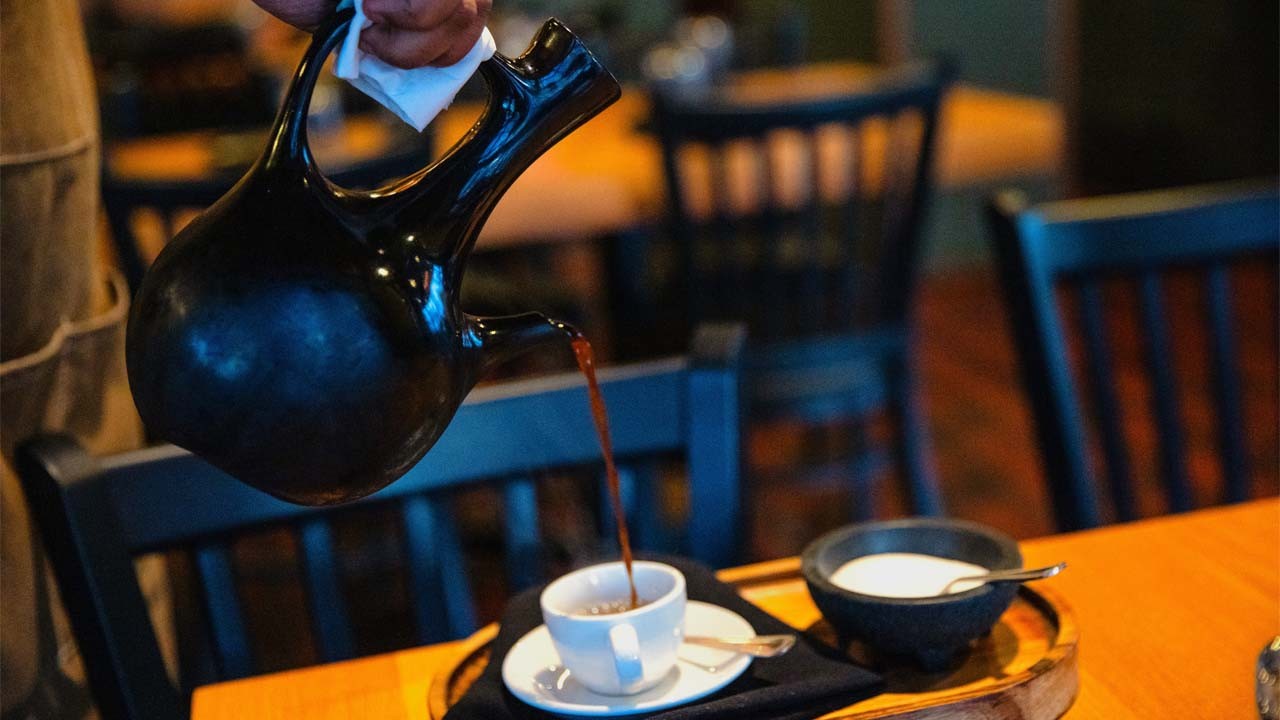 A barista pouring coffee from a black pot into a white cup, with a small bowl of cream on the side.