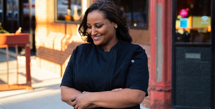 A smiling female chef in a black uniform standing outside a restaurant, with her arms crossed and looking down.