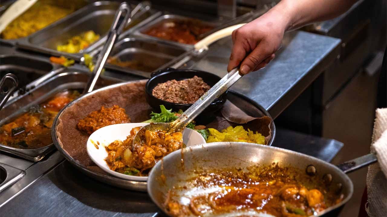 A chef using tongs to serve a spicy dish onto a plate, surrounded by various food containers and ingredients in a busy kitchen.
