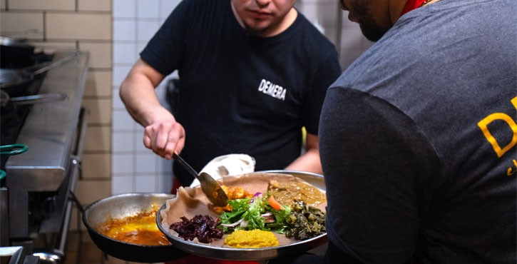 Two chefs preparing a colorful platter of food in a busy kitchen, with one chef scooping food and the other assisting.