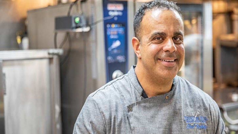 A smiling chef wearing a gray uniform, standing in a kitchen with Rational cooking equipment in the background.