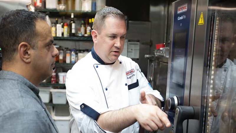 A chef in a white uniform explaining the features of a Rational cooking oven to another chef in a gray uniform.