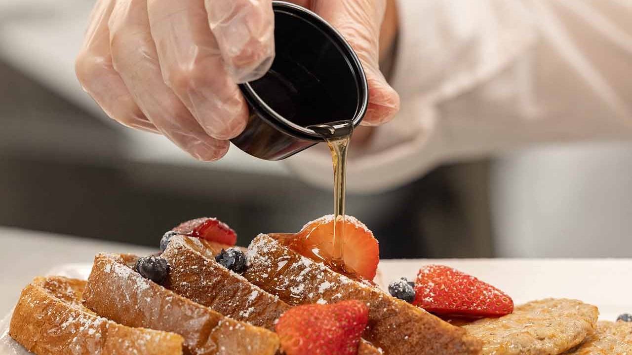Hand pouring syrup over a plate of French toast with strawberries and blueberries.