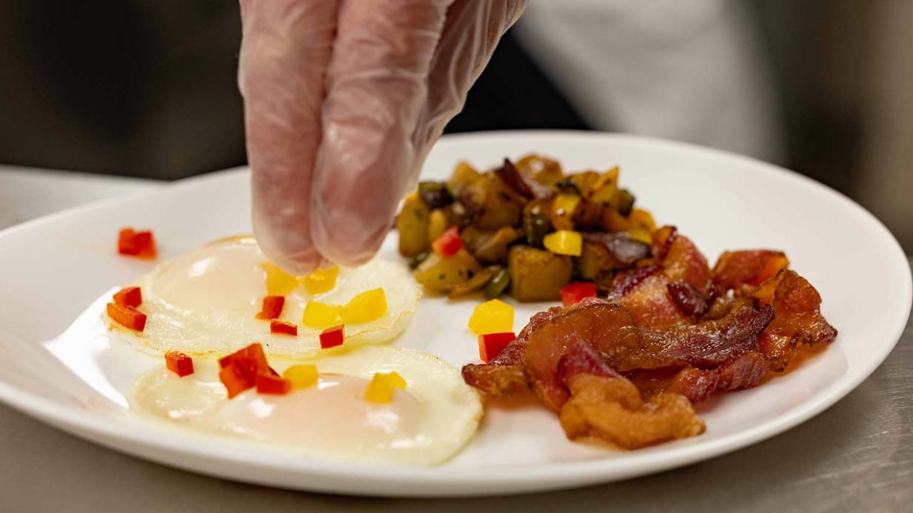 Hand sprinkling diced peppers on sunny-side-up eggs served with bacon and sautéed vegetables.
