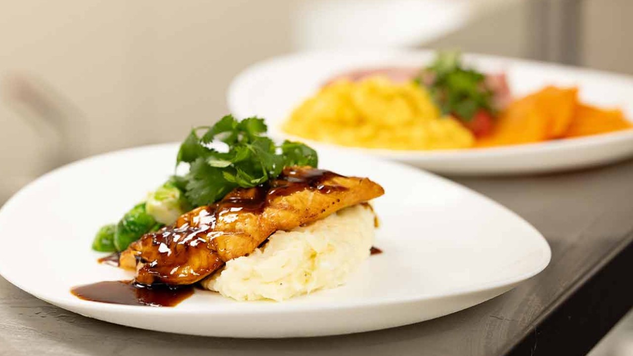Grilled salmon fillet drizzled with sauce, served on a bed of mashed potatoes and garnished with cilantro, accompanied by steamed vegetables.
