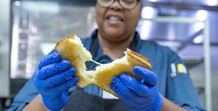 A chef holding a gooey grilled cheese sandwich, showcasing its melted cheese interior, with a smile in a kitchen setting.