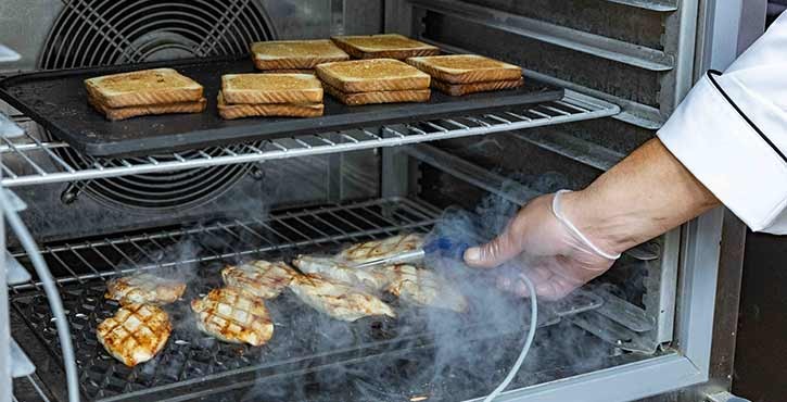 A chef‘s hand, wearing a glove, using a temperature probe to check grilled chicken while bread toasts on a rack in the iCombi oven.