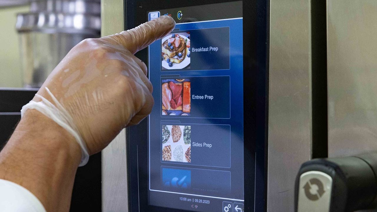 A chef‘s hand in a glove interacting with a digital control panel on an iCombi oven, selecting meal prep options.
