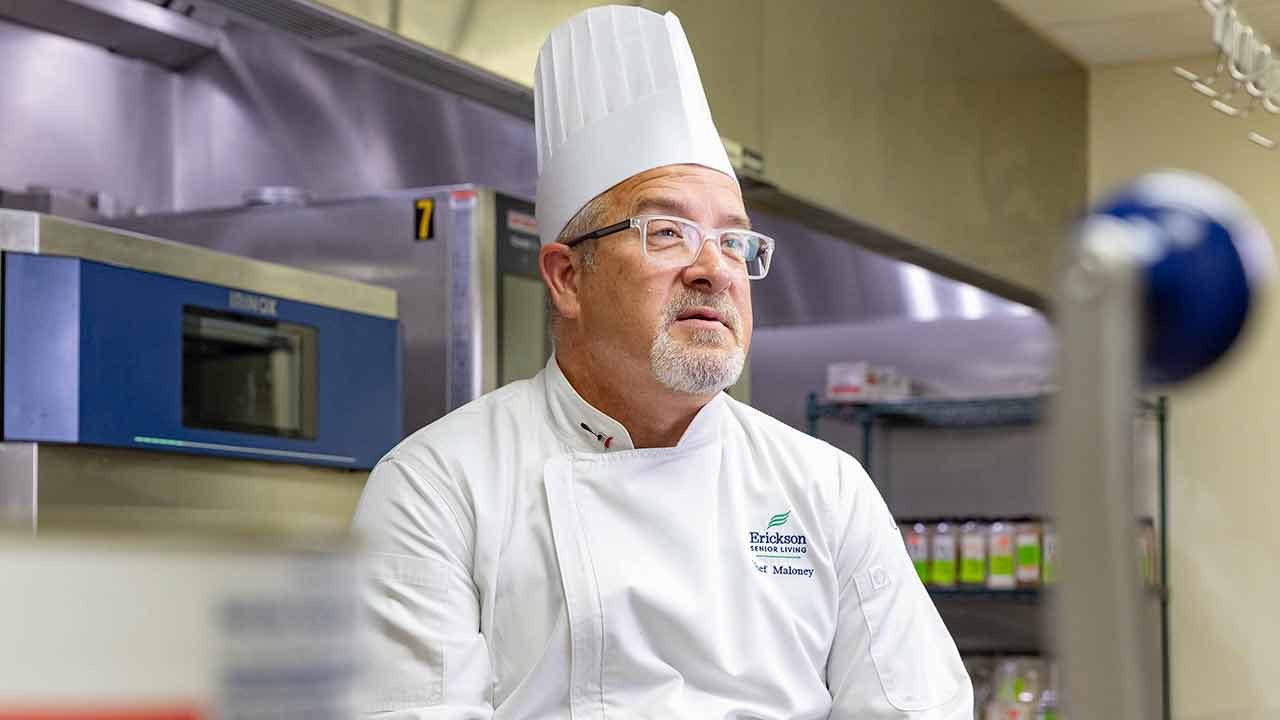A chef wearing a tall white hat and glasses sits in a kitchen, looking thoughtfully while surrounded by kitchen equipment.