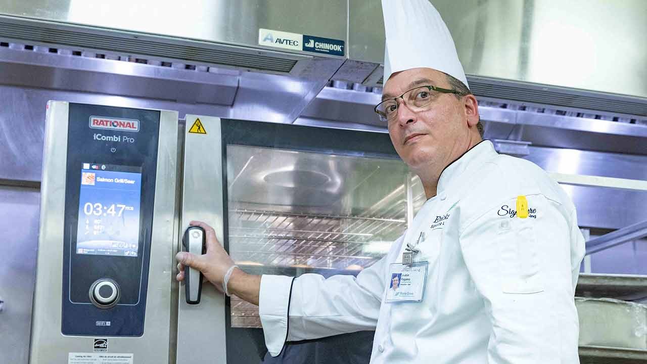 A chef in a tall white hat adjusts the control panel of an iCombi Pro oven while looking at the camera.