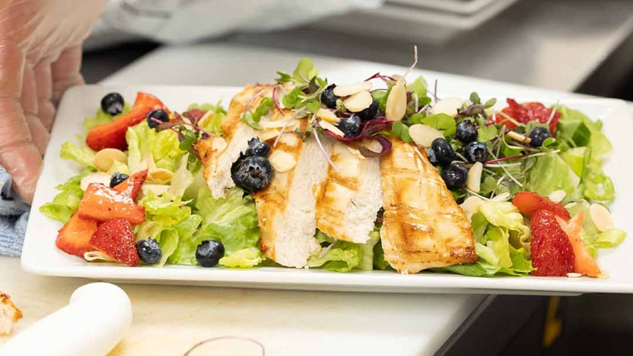 A hand in a glove places a grilled chicken salad topped with strawberries, blueberries, and almonds on a plate.