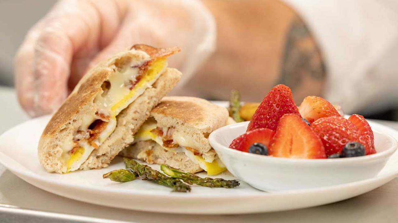 A hand in a glove holds a sandwich filled with eggs, cheese, and bacon, with a side of strawberries and asparagus on a plate.